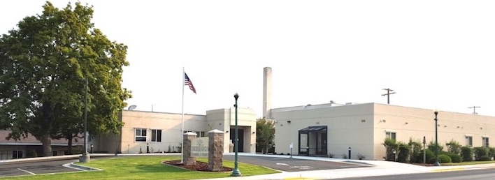 Picture of Syringa Hospital. There is a flag pole with the American Flag, a big tree, and parking spaces outside of the building